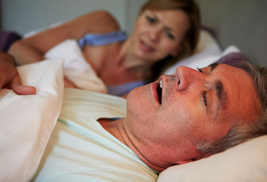 Man snoring in bed with an upset, awake wife in the background